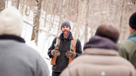 Man in orange safety vest speaks to people in the woods in winter