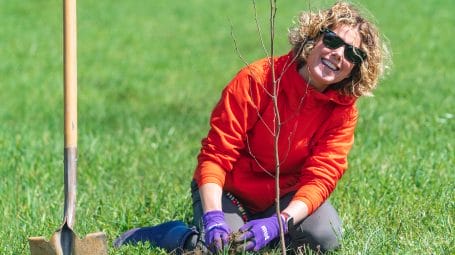 woman smiling at camera as she plants a tree in a hole she has dug - volunteers planted trees for clean water at a farm in Barre, Vermont