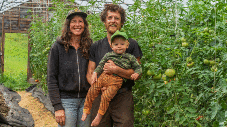 Marina, Andy and Cosmo stand in greenhouse with tall tomato plants at Heartwood Farm, where they practive sustainable agriculture