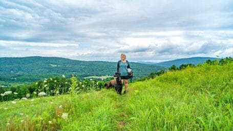 Woman with dog walking along hillside with valleys and hills in distance - Tracy Zschau - Vermont Land Trust