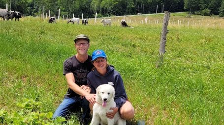 Couple sitting in farm field with white dog, with cows grazing in pasture behind them. Wild Earth Farm