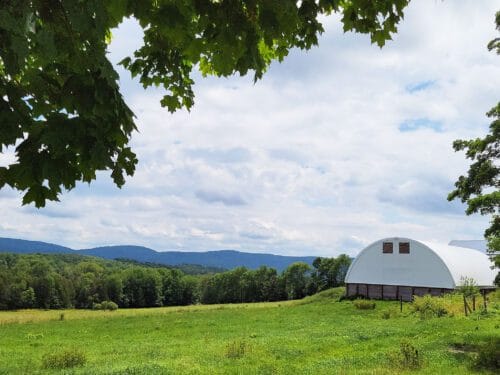 Open scenic view of farm pasture, white hooped barn to the right and distant trees and hills. Wild Earth Farm