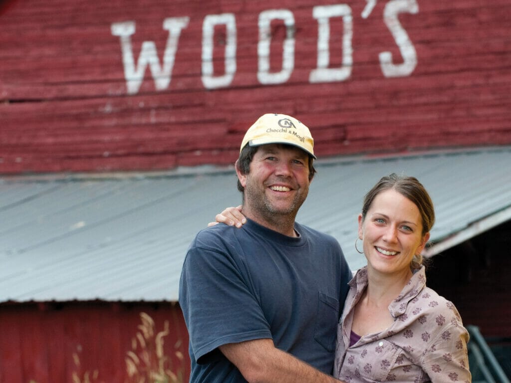 Man and woman smiling at camera with wooden barn behind them with 'Wood's' painted on it - Woods Market Garden - Jon and Courtney Satz