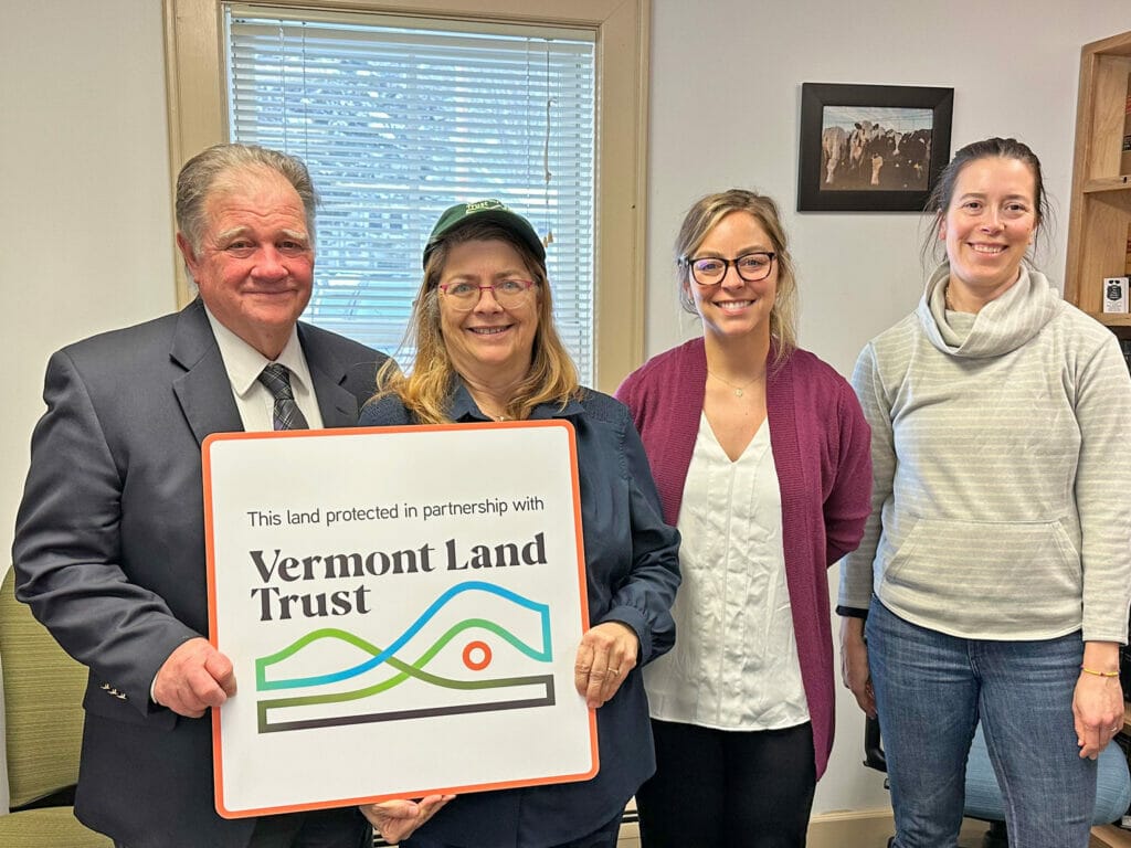 Four people standing in a room smiling at the camera; two holding a large sign that says Vermont Land Trust. Joe Tisbert and and Anne Marie Tisbert of Valley Dream Farm, with VLT staffers