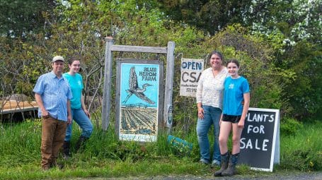 smiling family of four - man, woman, and two daughters - standing next to sign that says 'Blue Heron Farm' and 'Open CSA' and 'Plants for sale'