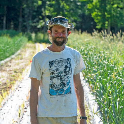 Man wearing hat standing in farm field with rows of corn behind him - Sam Rowley wins Land & Lives Award 