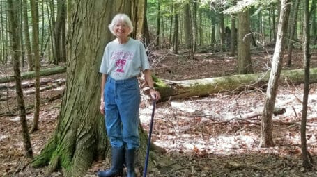 Smiling woman standing in front of wide-trunked tree in woods. South Burlington. Vermont.