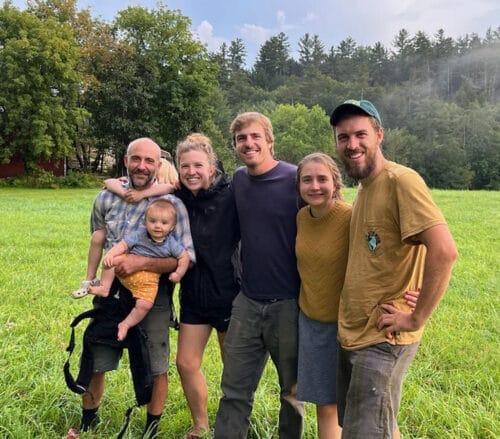 Five men and women standing close together, smiling, in a farm field with trees in distance. The Farm upstream collective, in Jericho Vermont