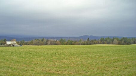 Fam field with trees and hills in distance and farmstead to the left - Marshfield farm, Vermont