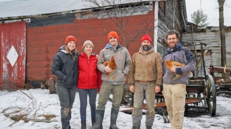 Group of five farmers -- two holding chickens -- standing in farmstead with barn behind them. The Farm Upstream. Jericho Vermont. collective farm
