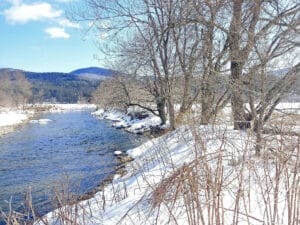 Sunny snowy day along a river with trees along bank, farm fields, and hills in distance. DeFreest Farm