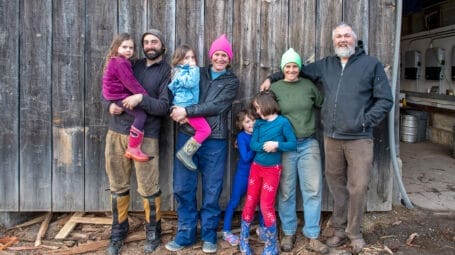 group photo of two farm families with children, standing in front of a gray barn. Bunker Farm Vermont