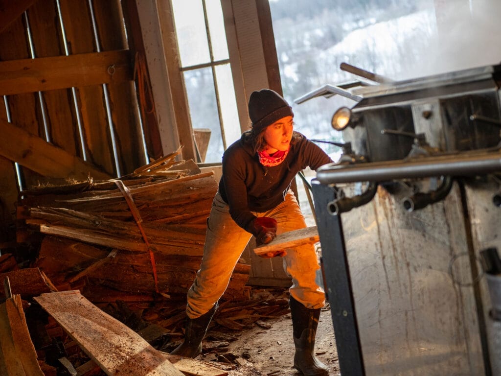 Woman loading firewood into large metal furnace for maple syrup evaporator. Bunker Farm Vermont