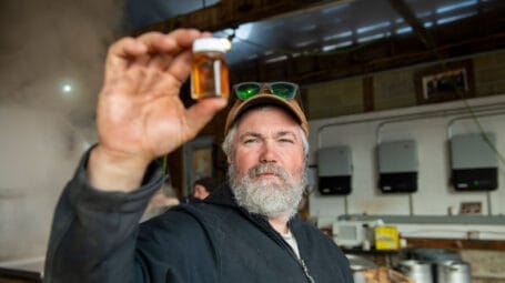 Close-up of man holding small glass jar with golden maple syrup inside - workshed in background. Bunker Farm, Dummerston Vermont