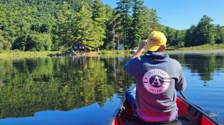 View from back of man paddling in canoe to small island with hut. Cam Dream, Vermont youth summer camp expands.